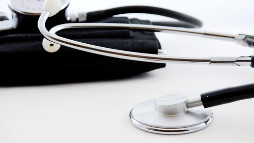 A stethoscope with a silver chest piece and eartubes rests on a white surface, near a blurred sphygmomanometer cuff.