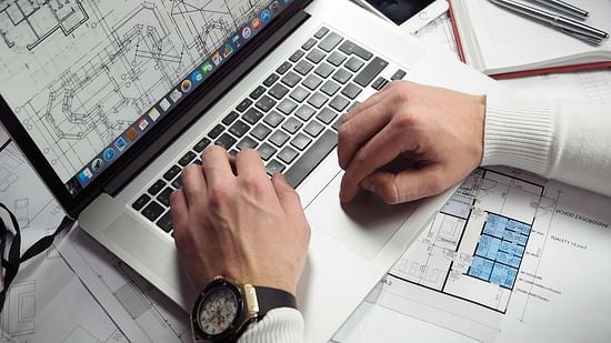 Hands of a man in formal attire working on a laptop, surrounded by papers.