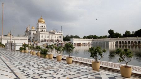 A white gurudwara with a golden dome beside a large sarovar, featuring a patterned marble courtyard, potted plants along the walkway, and visitors gathered near the water.