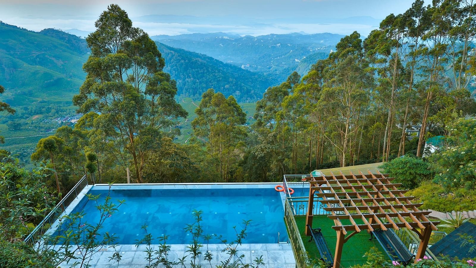 Aerial view of a swimming pool nestled in a lush green forest, with mountain ranges in the background – Fragrant Nature Munnar.