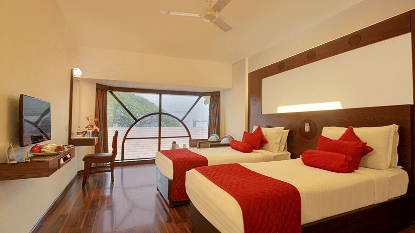 Executive Room at The Monarch Hotel, Brigade Road, Bangalore, with a bed, red cushions, a large window view, a ceiling fan and polished wooden flooring.