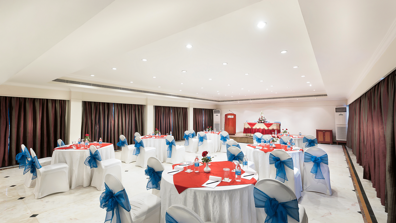 A banquet hall with white chairs laced with blue ribbons pointing towards the stage at Regency Kanchipuram by GRT Hotels.