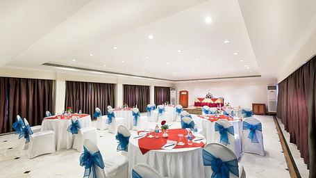 A banquet hall with white chairs laced with blue ribbons pointing towards the stage at Regency Kanchipuram by GRT Hotels.