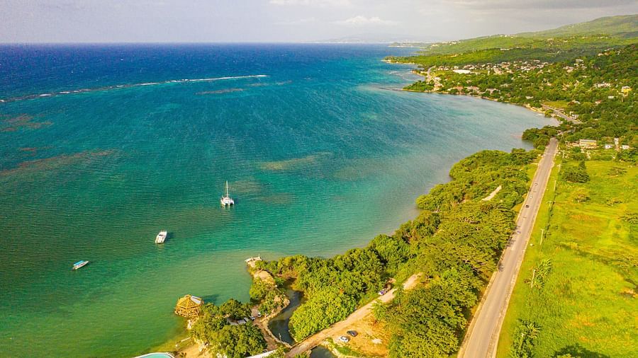 a vibrant coastline with turquoise waters, lush green land, a road, scattered buildings, and several boats.