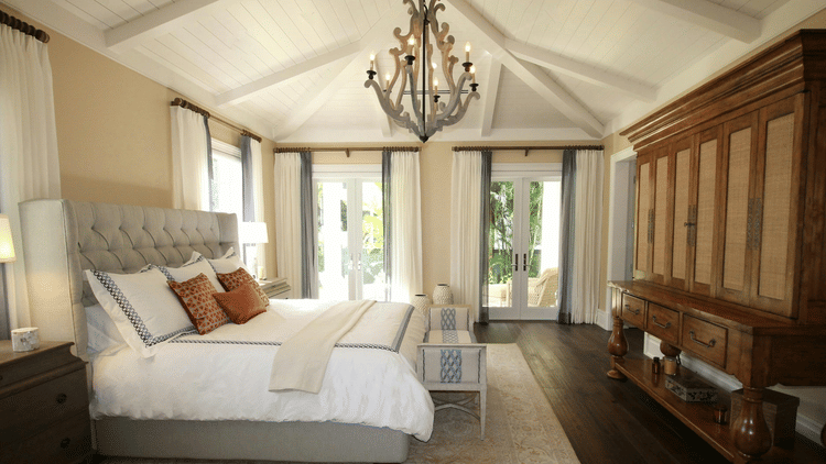A spacious, elegant bedroom with a tufted bed, wooden furniture, chandelier, large windows, and soft natural light creating a warm, airy ambience.