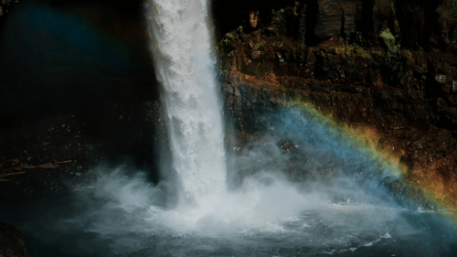 A far out view of a waterfall cascading down to a waterbody with a rainbow at the bottom right