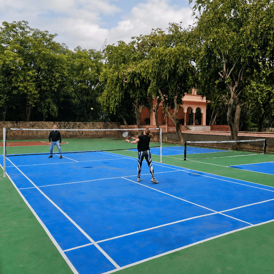 Two people playing on a bright blue outdoor badminton court surrounded by lush green trees and a gazebo under a cloudy sky.