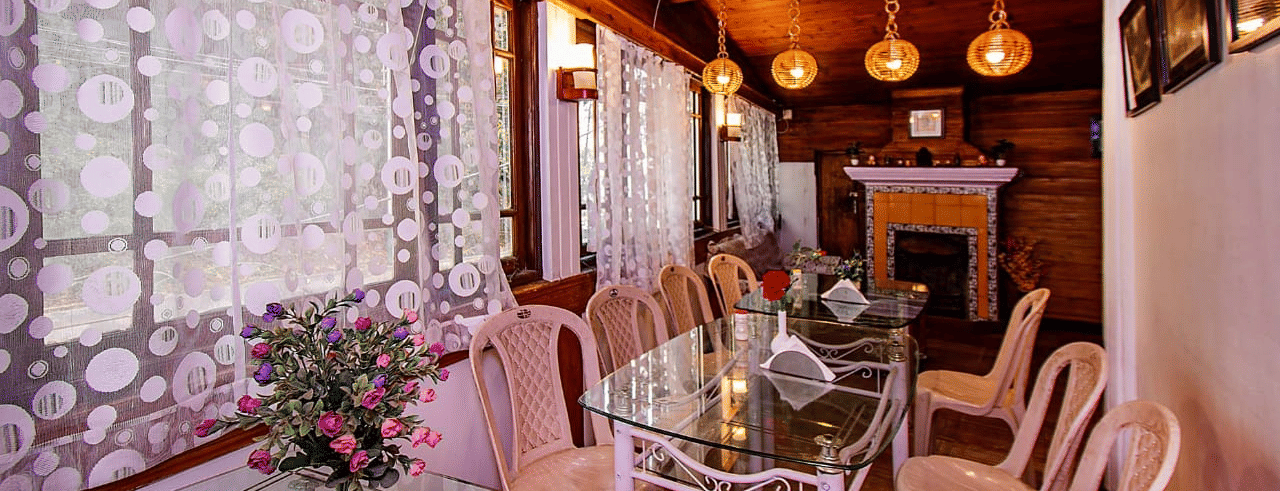 Dining area with a wooden ceiling, hanging lights, a glass table, and floral patterned white lace curtains at Hotel Dolphin Darjeeling.