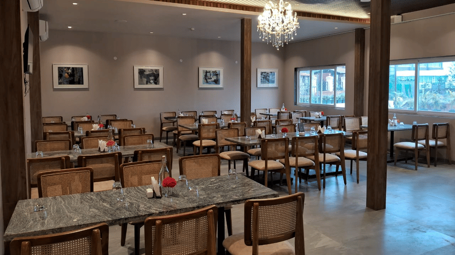 A spacious dining hall, filled with wooden tables and chairs under hanging chandelier lights at Savyasaachii Hotel.