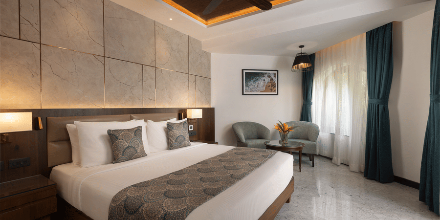 A view of the Junior Suite with a king bed, side tables, side lamps, ceiling fan and chairs in the corner at Kenilworth Resort & Spa, Goa.