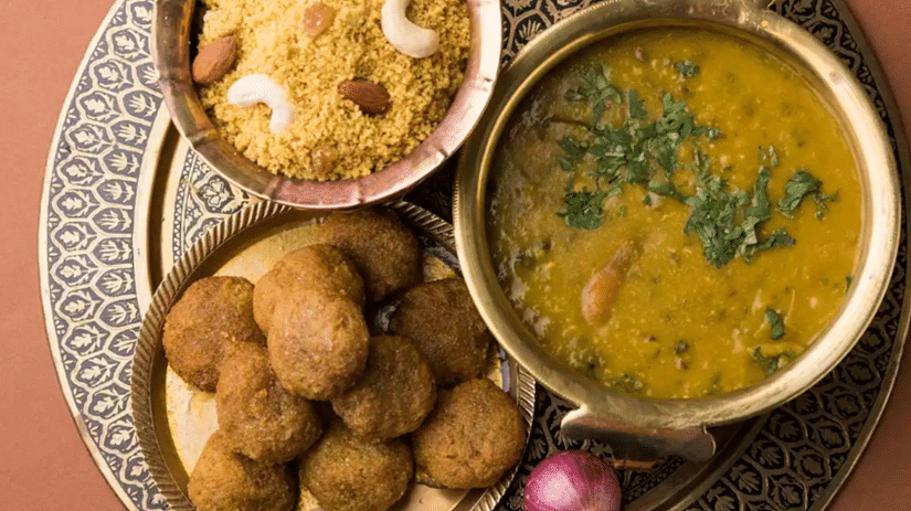 A top view of delicious Dal Bati Churma served on a plate, featuring baati, dal, and sweet churma, accompanied by shallot onions and green chilies for added flavour.