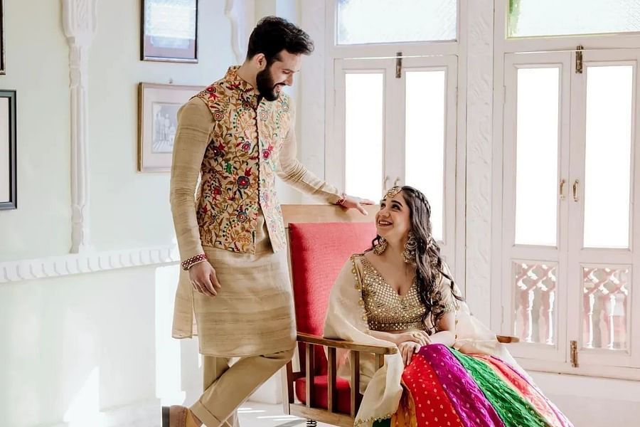 A couple smiles at each other, with the woman seated and the man standing beside her, in a warm setting- Umaid Palace - Best Luxury Heritage Resort Near Jaipur.  