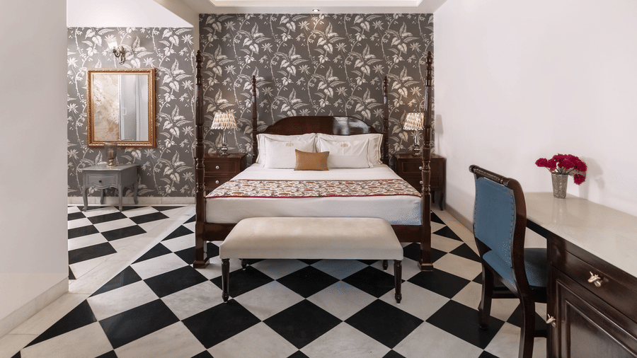 The Deluxe Heritage Suite in Jaipur at Dileep Kothi with a four-poster bed, a patterned wallpapered wall, a chequered black and white tiled floor, and a writing desk with a blue chair.