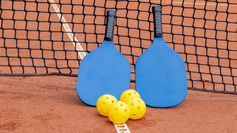 Two blue pickleball paddles leaning against a net with yellow pickleballs on a court surface.