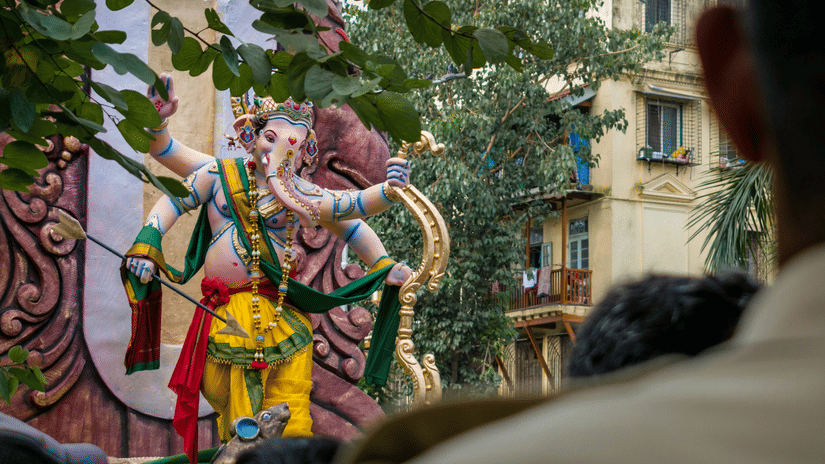 A large colorfully decorated Lord Ganesha idol during an outdoor procession with trees and buildings visible in the background and crowd of people gathered in the foreground.