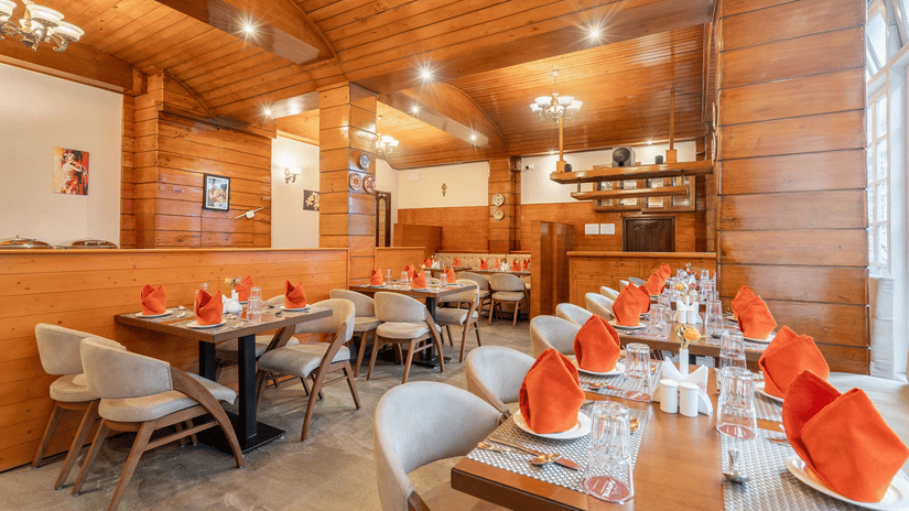 An indoor restaurant dining area with wooden tables and chairs, featuring tables set with orange fabric napkins.