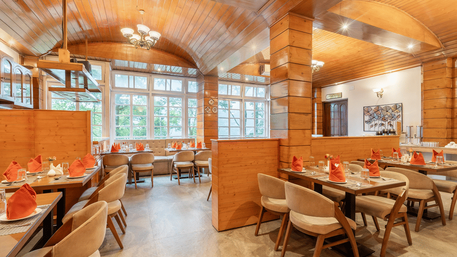 A brightly lit hotel bedroom featuring a bed with crisp white linens and a patterned runner, set against warm wooden wall panelling with a large flat-screen television mounted above a built-in dressing table.