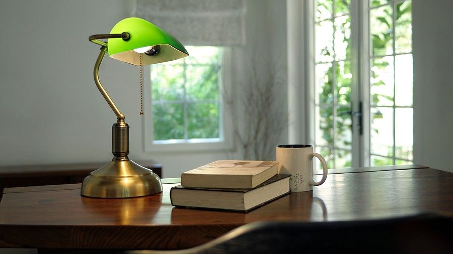 Work desk with a green lamp, books, and a mug by a window with sheer curtains.