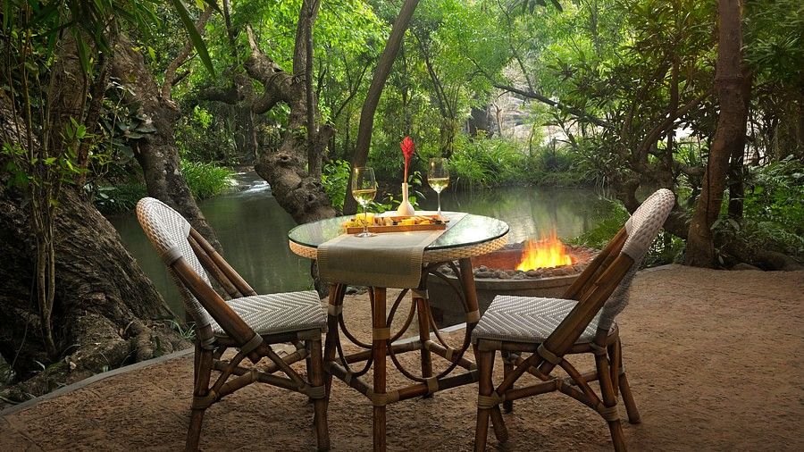 Riverside dining setup with lit candles and comfortable chairs, surrounded by trees.
