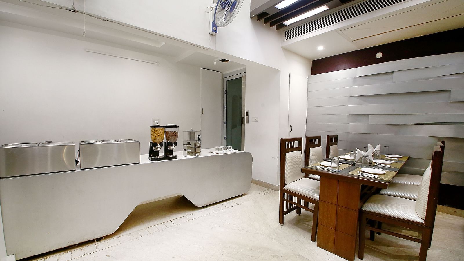 Dining space with a table and four chairs around it in a corner, a table with white plates and a desk with utensils along with two fans in a modern interior setting - Hotel Emerald, Chandigarh