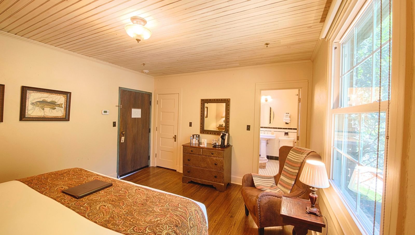 a cozy bedroom with a cushioned rustic armchair along with a small side table with a table lamp and a double bed in the room - Historic Tapoco Lodge