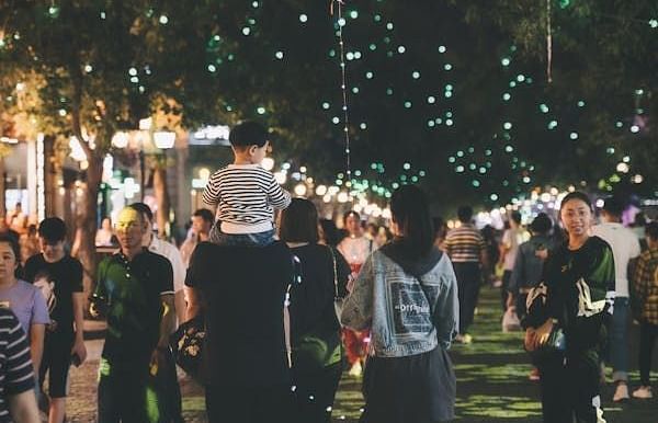 A person carries a child on their shoulders through a crowd, with lights strung overhead and a large, clear balloon visible.