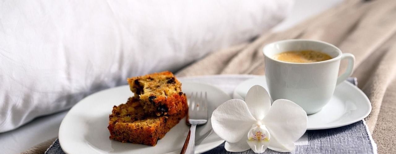 Plate with slices of cake and fork, cup of coffee, and white flower placed on a tray on a bed.