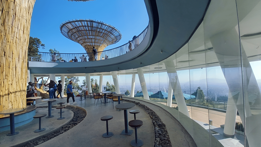 Modern architectural space with curved glass walls, round tables, people walking on upper level, and large tree?like lattice sculptures against clear blue sky.