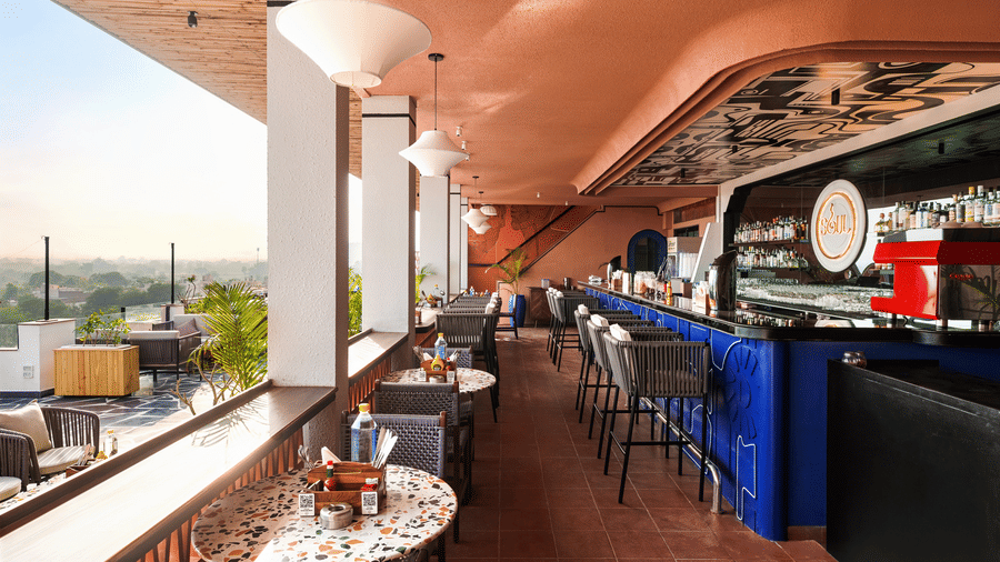 Stools line a vibrant bar counter on a rooftop patio at The Sierra - By The Lake, showing a view of the surrounding landscape past the open railing.