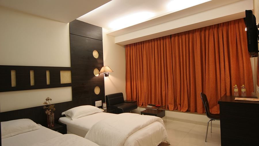 Business Rooms at Pride Biznotel Alkapuri featuring twin beds placed in front of the TV mounted on the wall and a work desk next to the window behind which a sitting area is organised 
