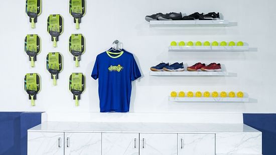 Display wall at SMASH Pickleball featuring pickleball paddles, a branded blue T-shirt, athletic shoes, and yellow pickleballs neatly arranged on white marble shelves at Jenneys Residency