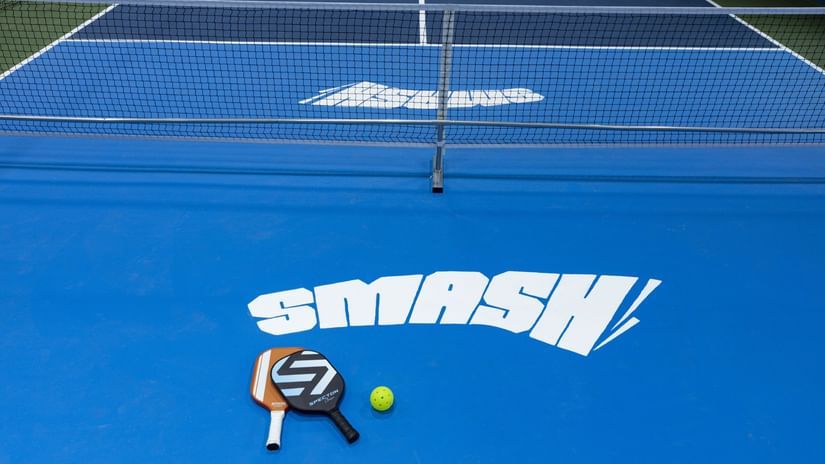 Close-up view of indoor pickleball courts with paddles and balls placed on a blue surface marked 'SMASH,' at Jenneys Residency, Coimbatore.