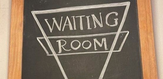A chalkboard sign with the text Welcome! WAITING ROOM and an arrow, next to a vase of sunflowers.