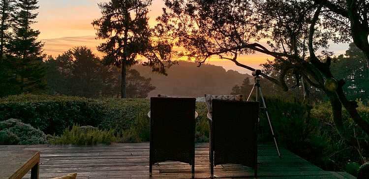 An image of a deck where two chairs are facing towards the sun set and the entire area is surrounded by trees