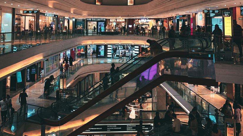 Multi-level shopping mall interior with an ornate domed ceiling, escalators, illuminated storefronts, and shoppers walking through the atrium.