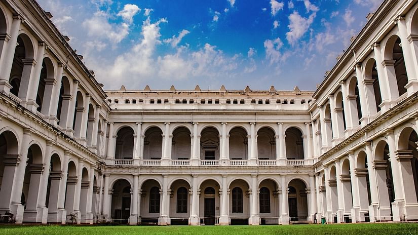 A grand white colonial-style building with arched corridors surrounding a green courtyard under a vibrant blue sky with scattered clouds.