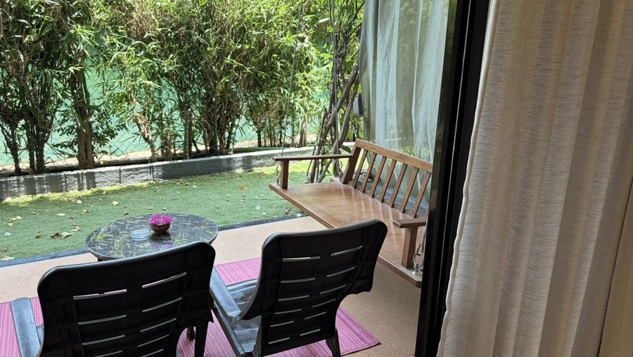 2 outdoor chairs and a coffee table rest on a pink mat next to a wooden swing overlooking a green garden hedge at Bougainvillea, Alibaug.