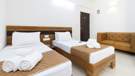 Twin bedroom with two beds, white linen, a sofa, and a wooden door at Sanctum Suites Whitefield, Bangalore.