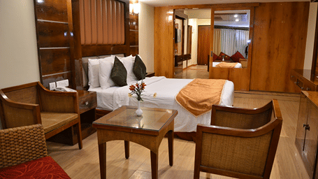 Room at Benzz Park with a wooden bed, green cushions, a seating area with cane chairs, a wooden coffee table and a view toward an adjoining dressing area.