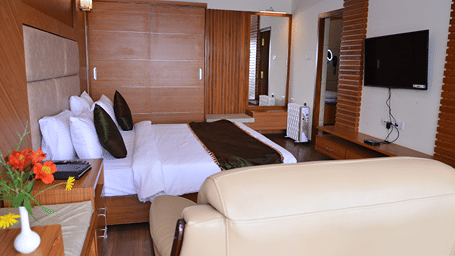 Room at Benzz Park with a wooden headboard, black cushions, a wall mounted television, a tray table, a cream sofa and fresh flowers on a side table nearby.