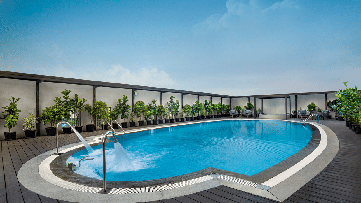An outdoor swimming pool at The Residency Towers Chennai surrounded by a timber deck and potted plants, with sun loungers visible