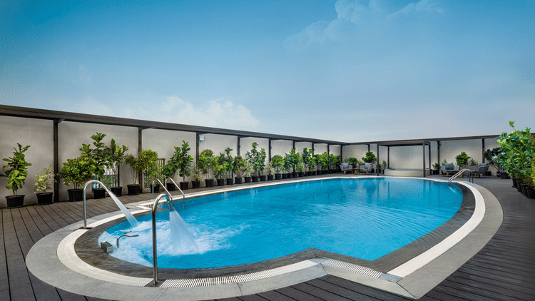 An outdoor swimming pool at The Residency Towers Chennai surrounded by a timber deck and potted plants, with sun loungers visible