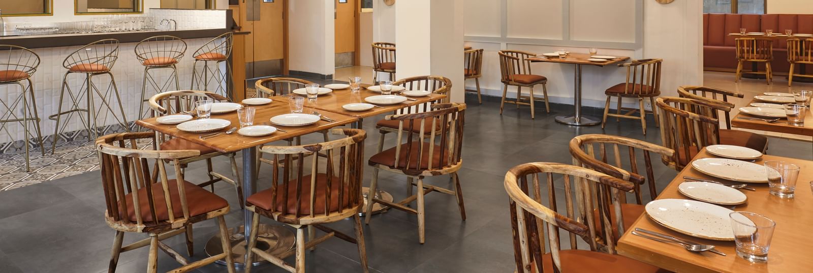 The spacious dining hall at Monday Hotels Mahape, Navi Mumbai, with multiple wooden tables and a central bar.