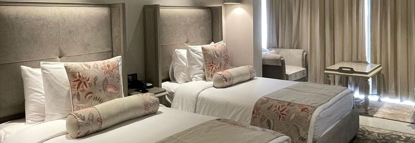 A spacious Premium Twin Room at The Evren, Vagator, with two single beds, each with a plush headboard and decorative pillows. The room features a patterned rug and a seating area near the window.