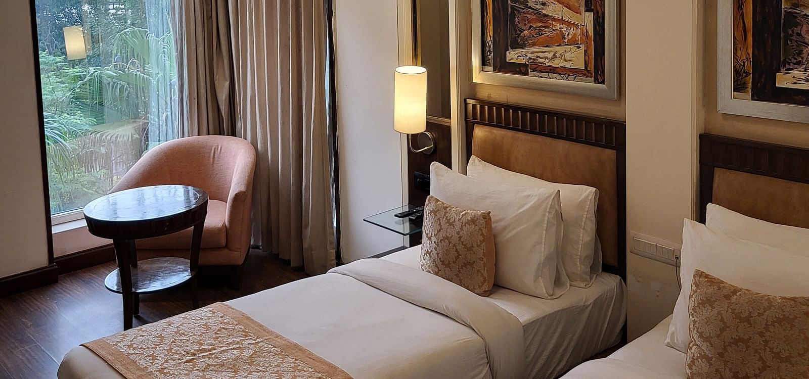 An image of a bedroom featuring twin bed, a sofa chair, a small centre table, bedside table with lamp, photo frames hanged on the wall, and a window o=overlooking the outdoors - Inde Hotel Signature Tower Gurgaon