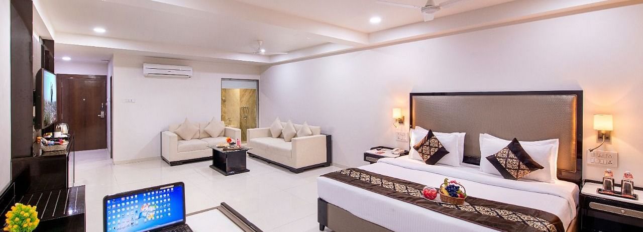 A comfortable hotel suite offers a king size bed, a separate seating area, a functional desk with a laptop, and ample natural light.