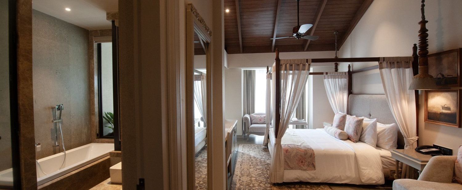 An elegant and spacious executive room at The Evren, Vagator, showing a four-poster bed with white curtains, a sloped wooden ceiling with a fan, and an open view into the luxurious bathroom with a bathtub.