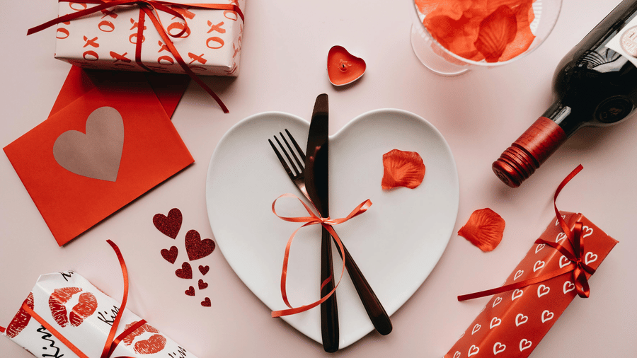 An image of a platter with cutlery tied with a ribbon, and around that there are heart shaped cards, gifts, and a wine bottle meant for valentine's day celebration