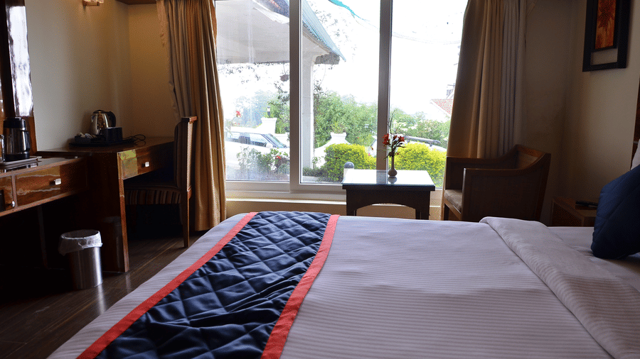 A room with a comfy bed, desk area, and balcony offering views of lush greenery - Benzz Park, Kodaikanal