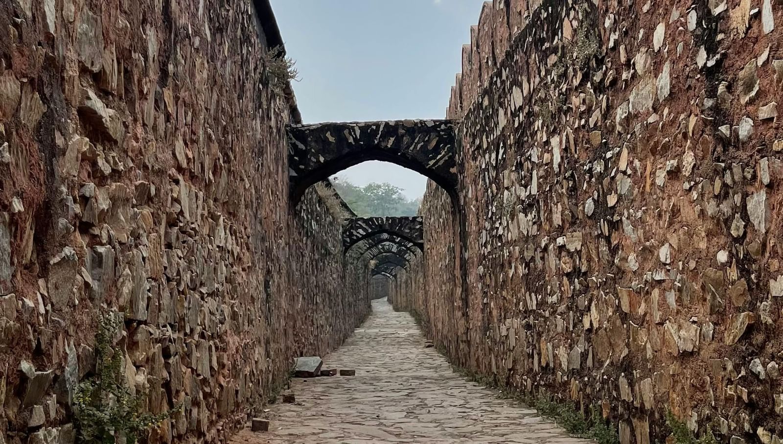 A narrow stone pathway between two high stone walls leading towards a small arched opening.